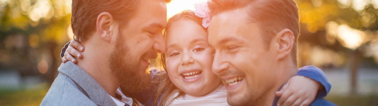 Young gay parents with their daughter having fun in park. Parents holding girl in arms. Enjoying in beautiful sunset.
