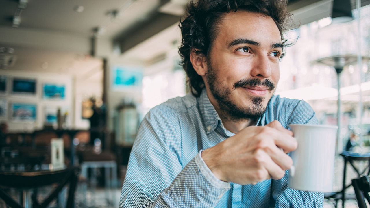 Young man sitting in cafeteria holding a cup of coffee and looking away, with copy space