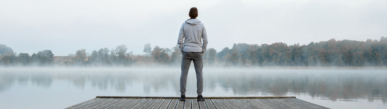 Young man standing alone on wooden footbridge and staring at lake. Thinking about life. Mist over water. Foggy air. Early chilly morning. Peaceful atmosphere in nature. Enjoying fresh air. Back view