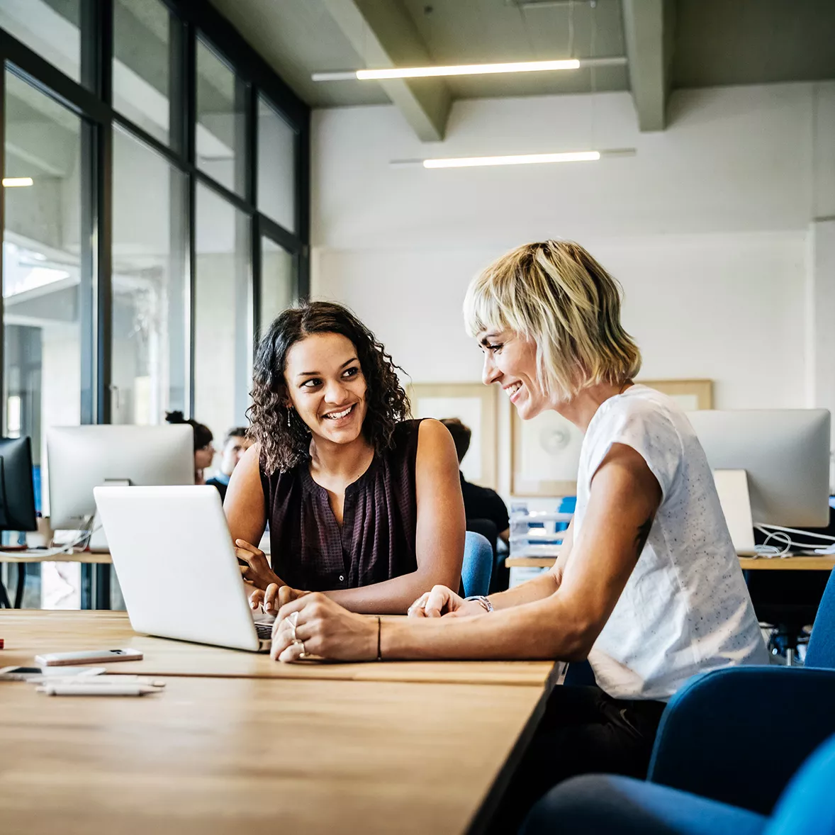 Two women sit at a table together, one providing customer support to the other on her computer