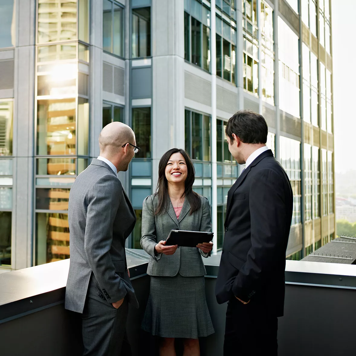 Three partners of different technology companies stand on a balcony together and review data 