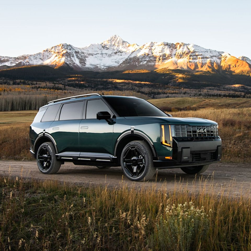 A still image of the 2027 Kia Telluride in blue-green parked on a dirt path, highlighting the passenger side of the SUV and the exclusive wheels, set against lush plants and snow-covered mountains.