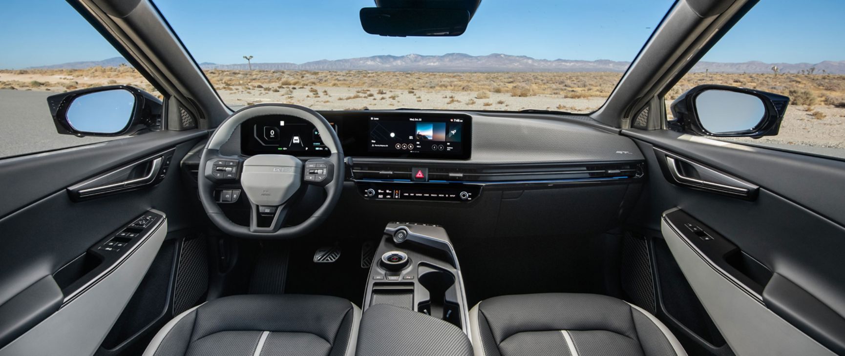 An interior front view of the 2026 Kia EV6, with a desert view and distant mountain range outside of the windows and windshield. The car's interior features a steering wheel, 12.3 inch dual panoramic displays, the dashboard, rearview mirrors, and more.