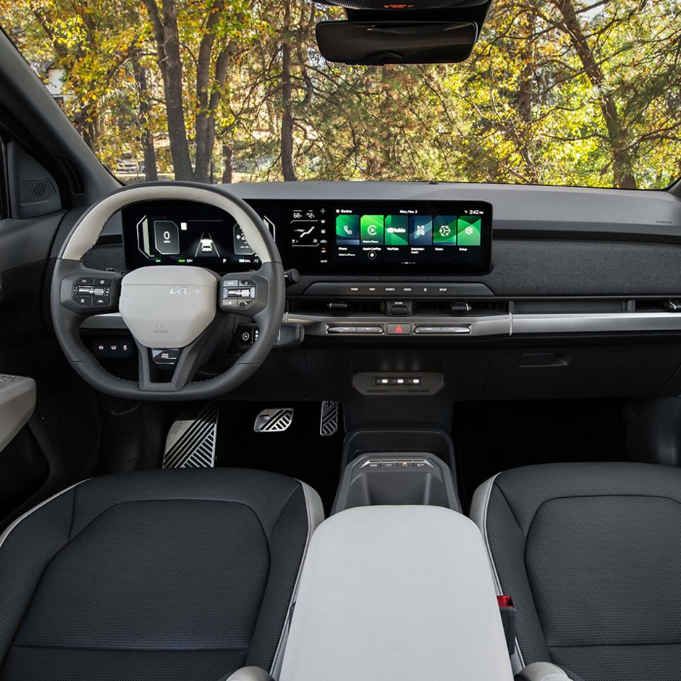 Interior wide-angle view of the 2027 Kia EV3 electric SUV showing seats, pedals, steering wheel, digital display, and more, with trees visible out the windows and windshield.