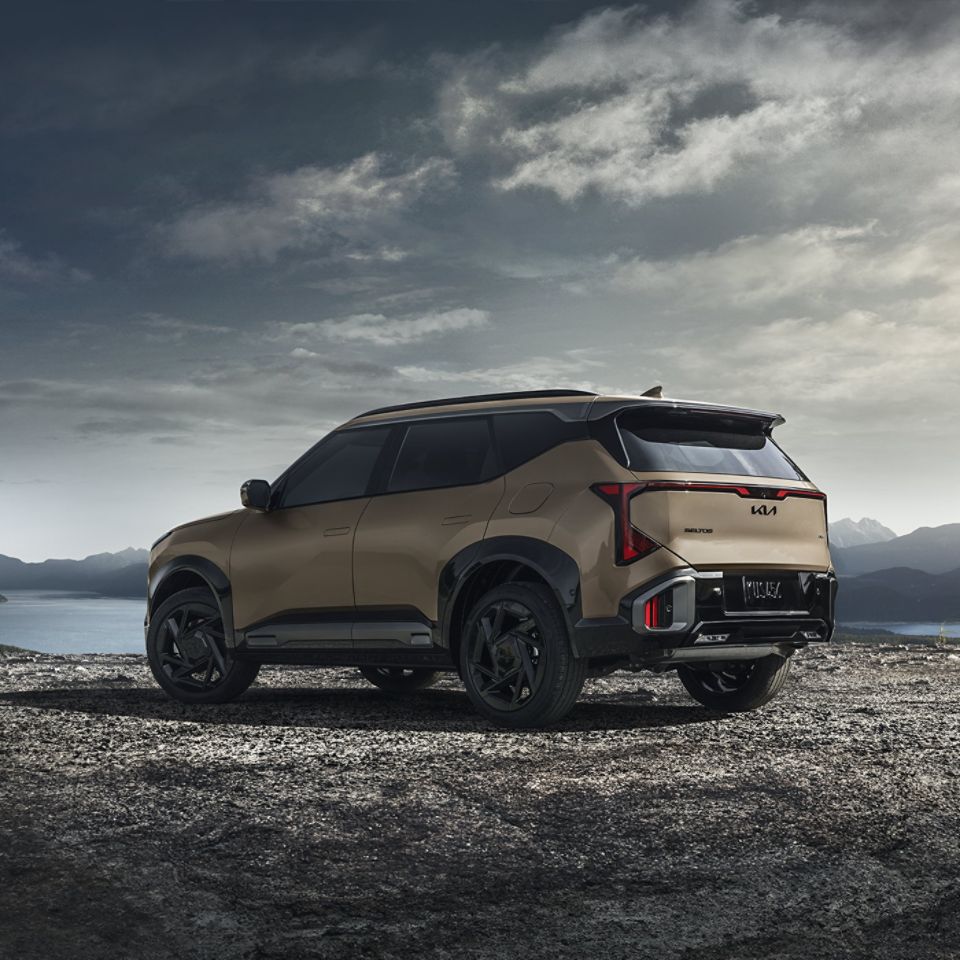 Rear-side photograph of the 2027 Kia Seltos SUV in tan as it is parked on a rocky road, with mountains and clody skies behind it. This photo emphasizes the elevated exterior design and the LED taillights.