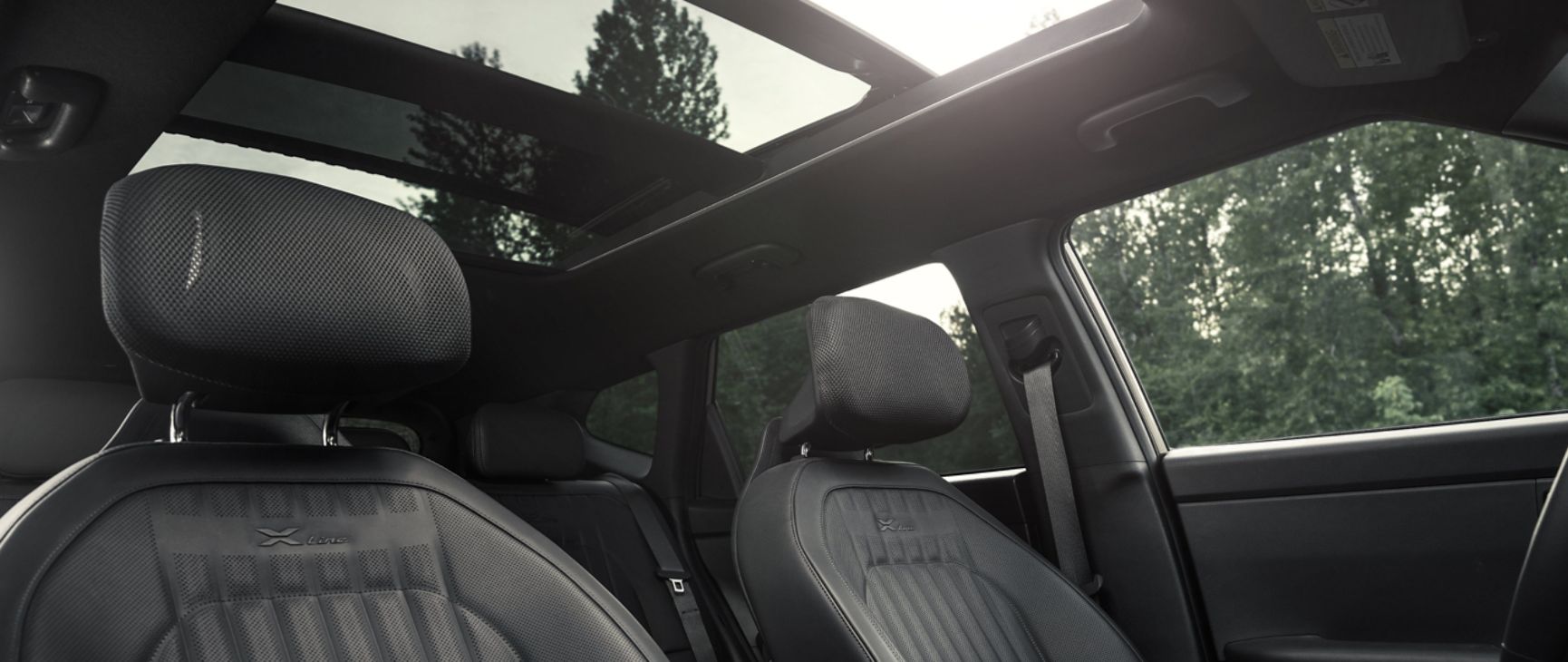Upward-angled interior photograph inside the 2027 Kia Seltos SUV showing its leather seats, comfortable headrests, and available panoramic sunroof, with tree visible and sunshine above the vehicle.