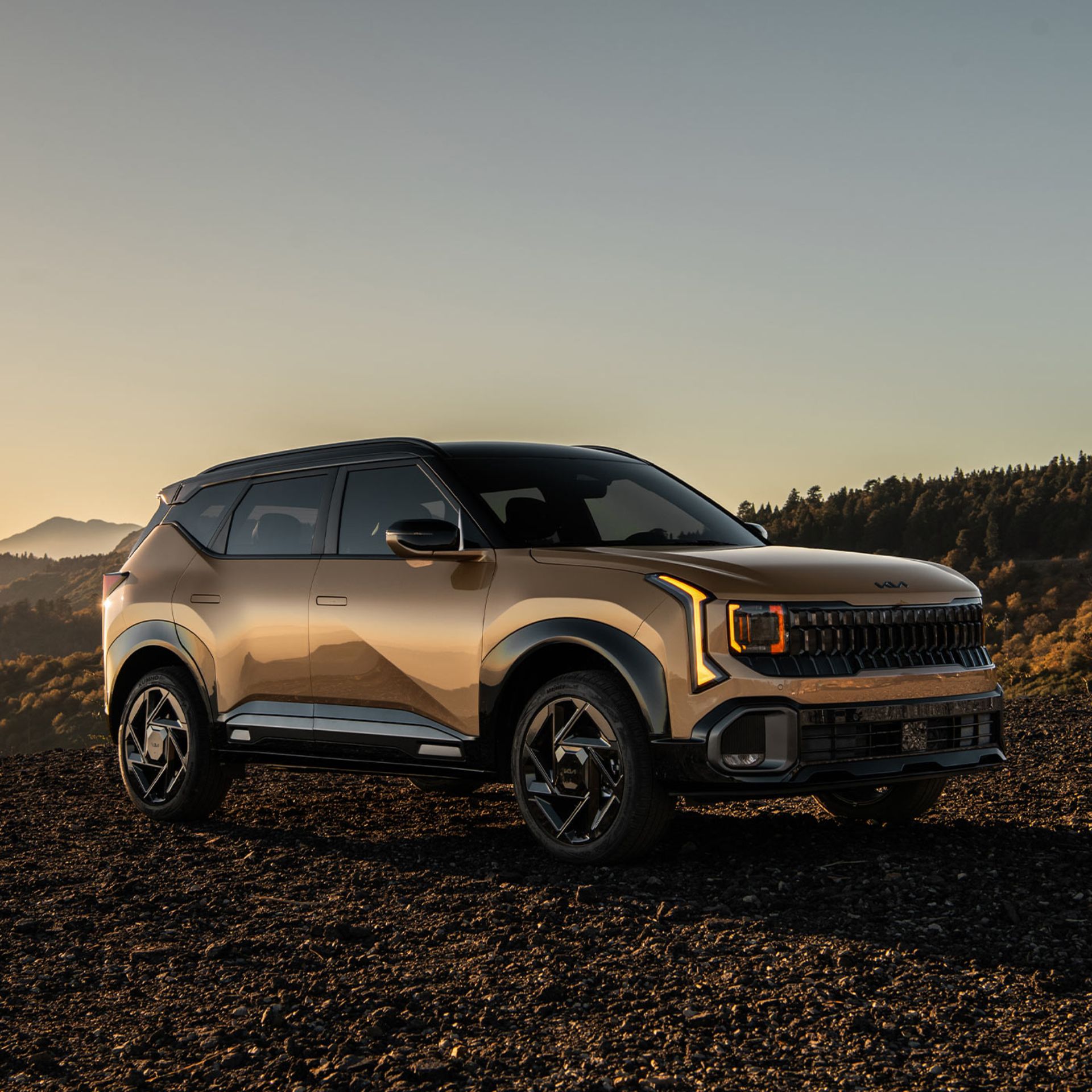 2027 Kia Seltos in brown parked outdoors at sunset, shown in a front three-quarter view with gloss black exterior trim, black side mirrors, and rugged styling against a mountain landscape.