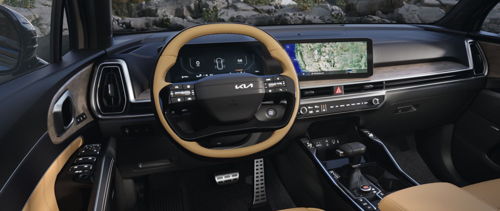 Interior wide-angle photo of the 2027 Kia Sorento in black and tan, showcasing its steering wheel, dual panoramic display with maps showing, center console, shifter, glove compartment, and more. A snow-filled forest is visible through the windshield.