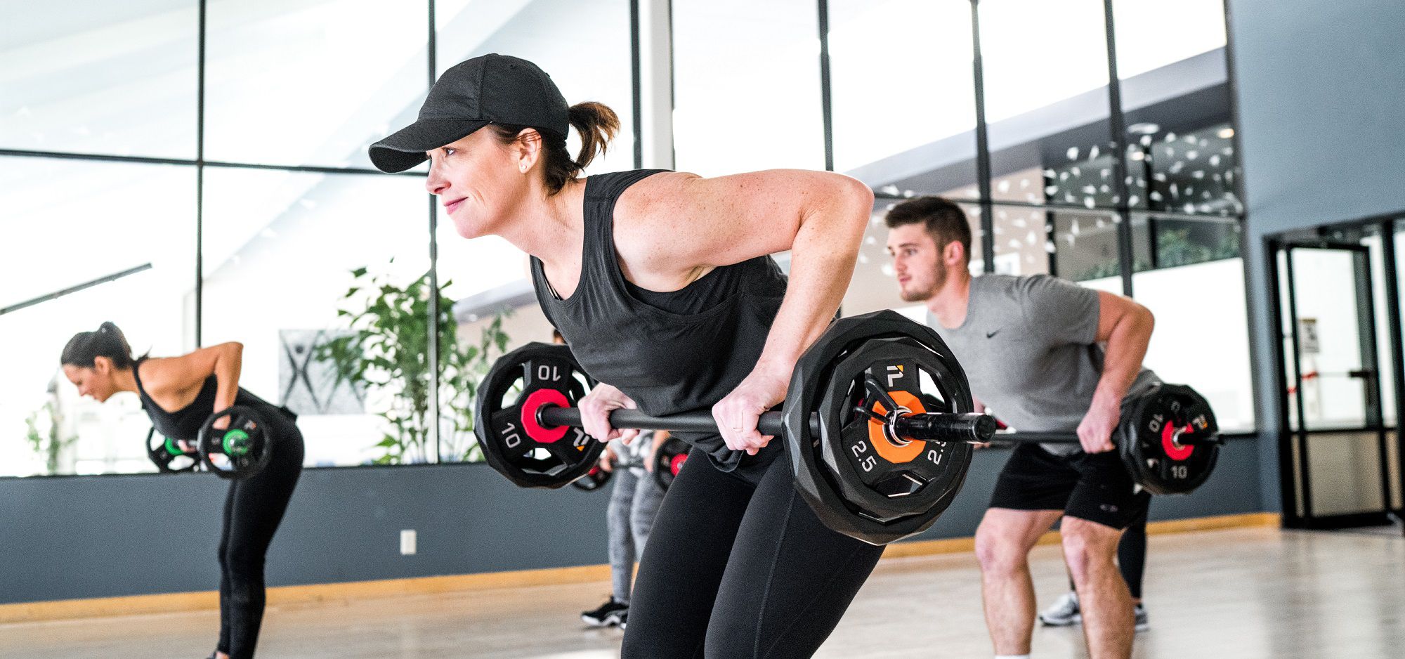 woman in a hat lifting a barbell with weights and other people in the background doing the same