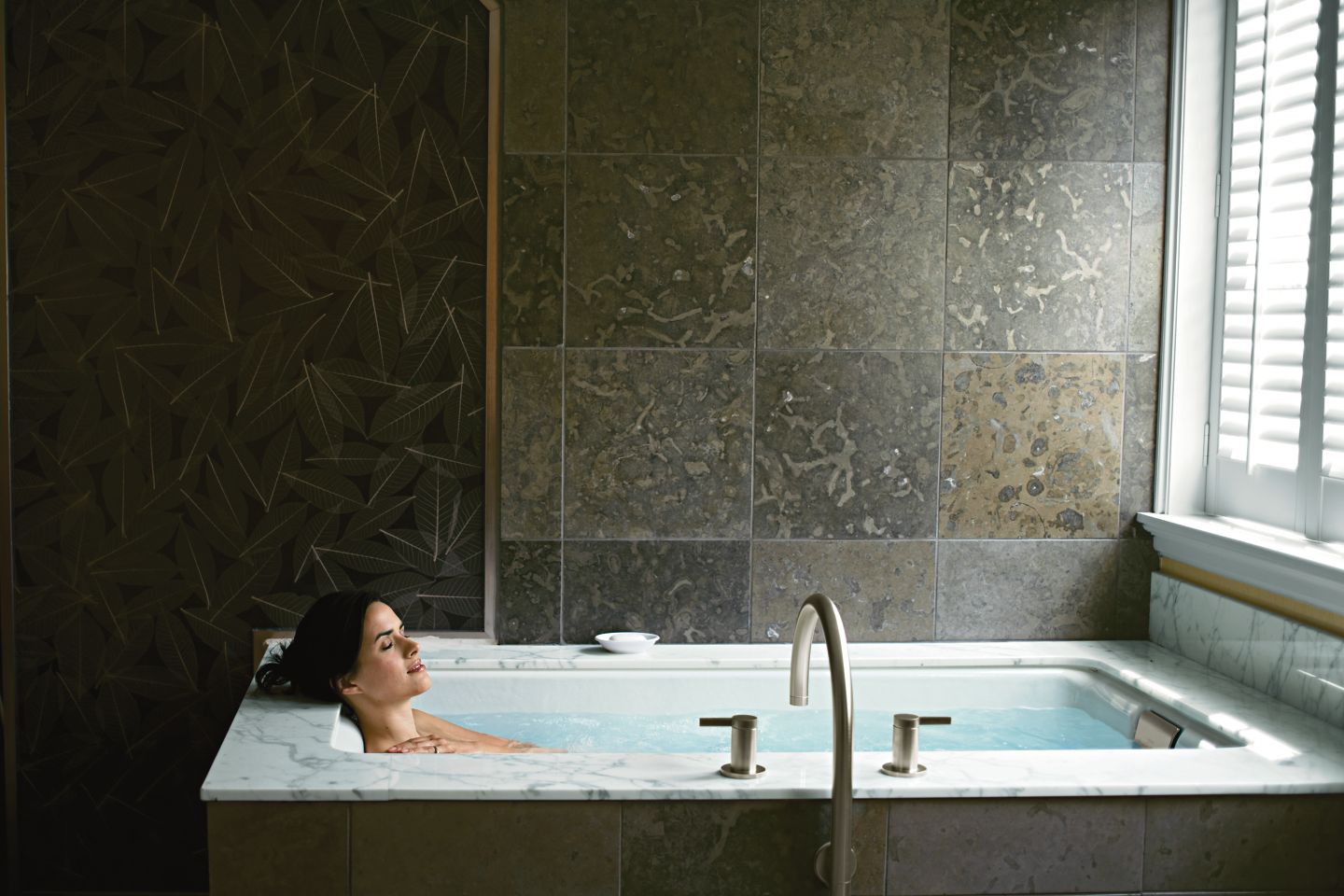 A woman relaxes in a Kohler bathtub facing a window
