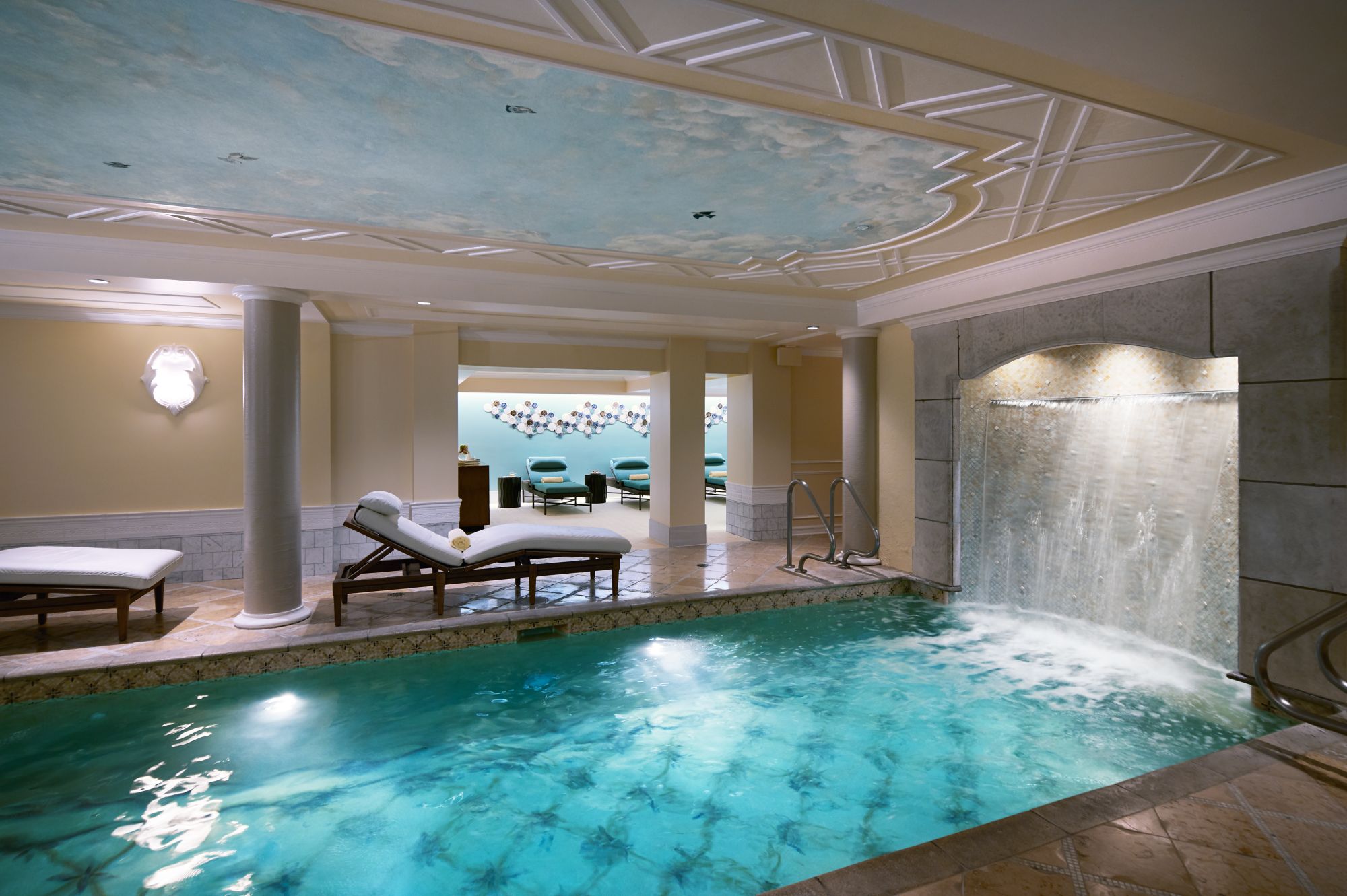 an indoor pool with a waterfall from the side view that focuses on another room with chairs
