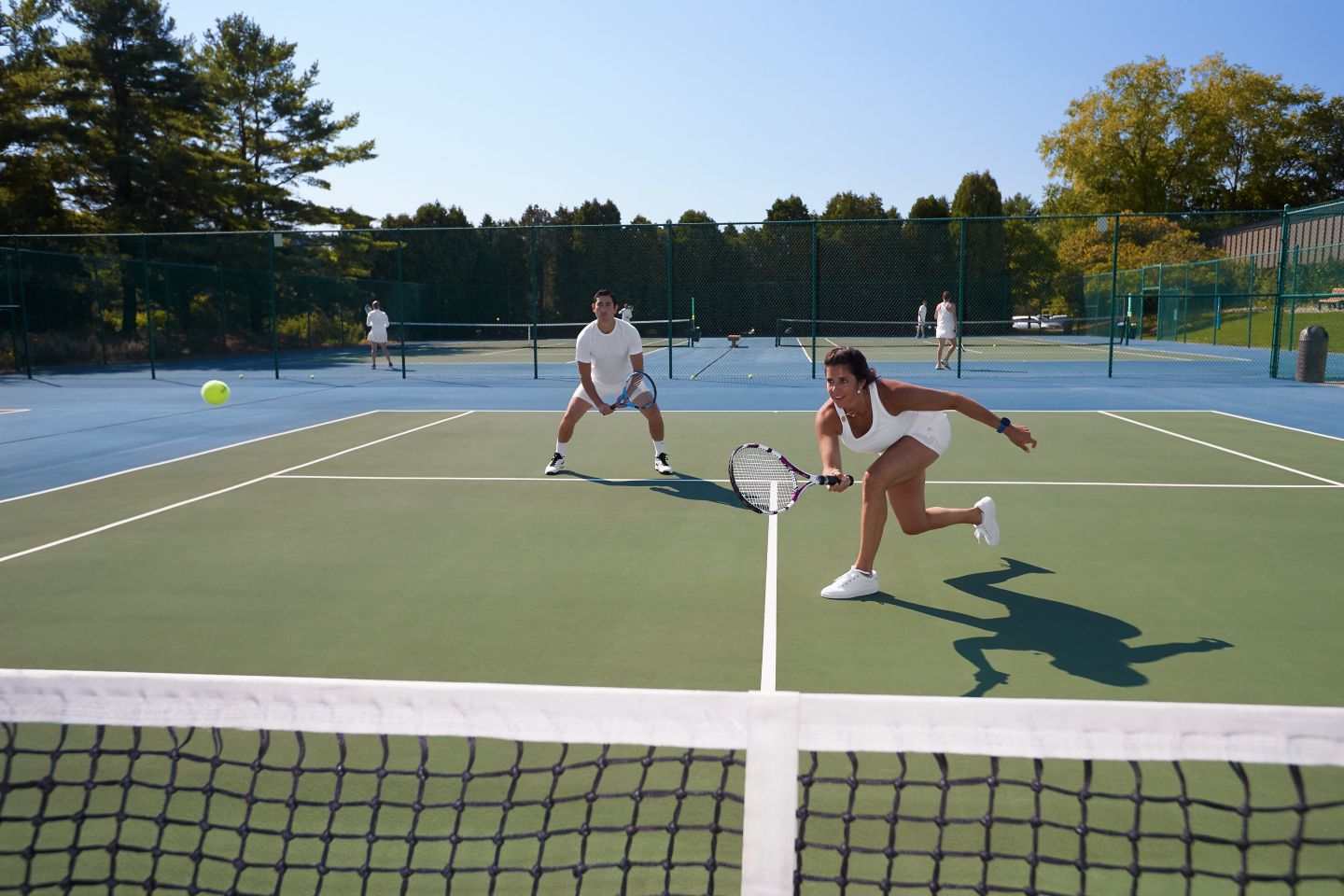 a lady hitting the ball over the net in a mixed doubles tennis match on an outdoor court