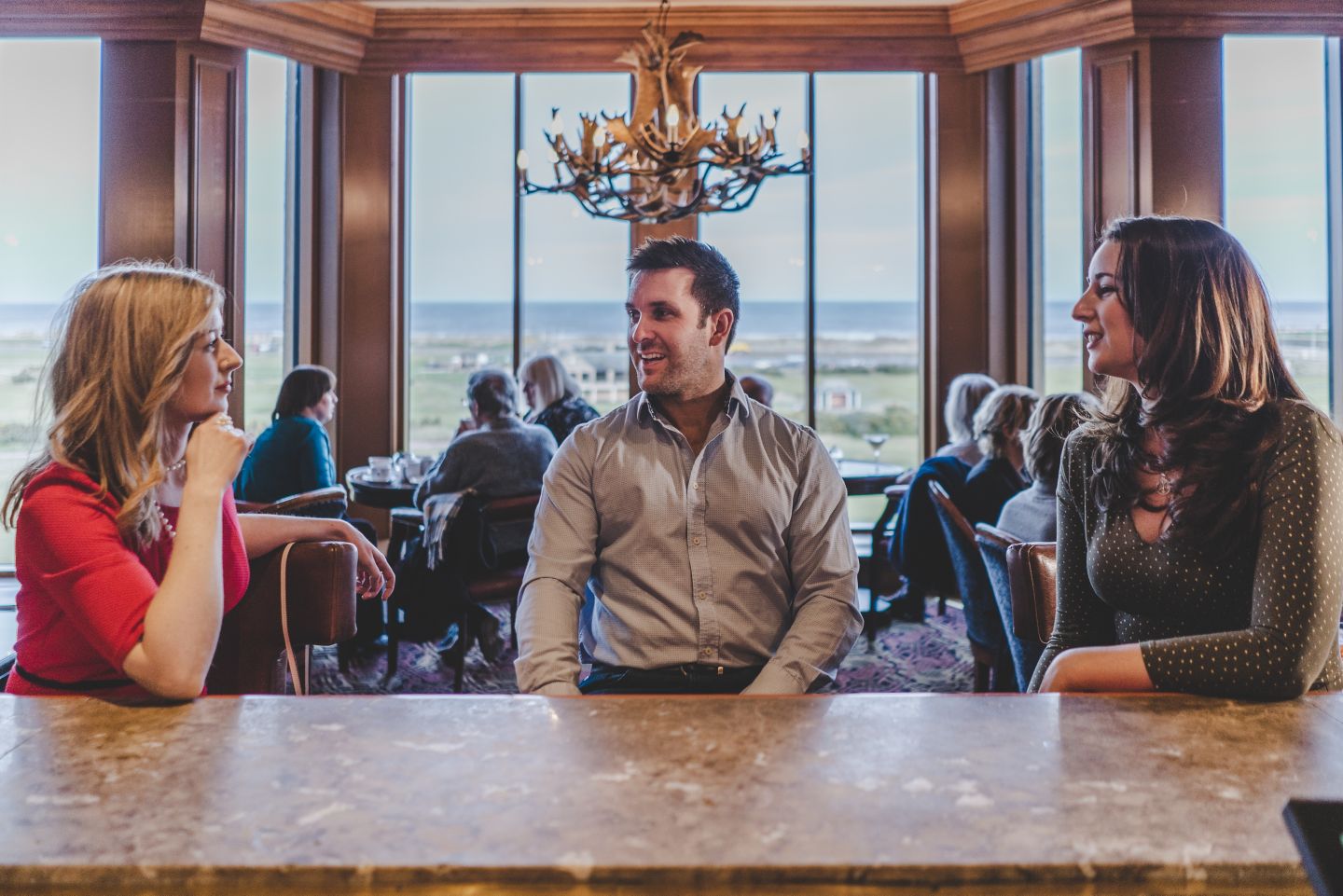 three people sitting at a bar with windows in the background overlooking the old course