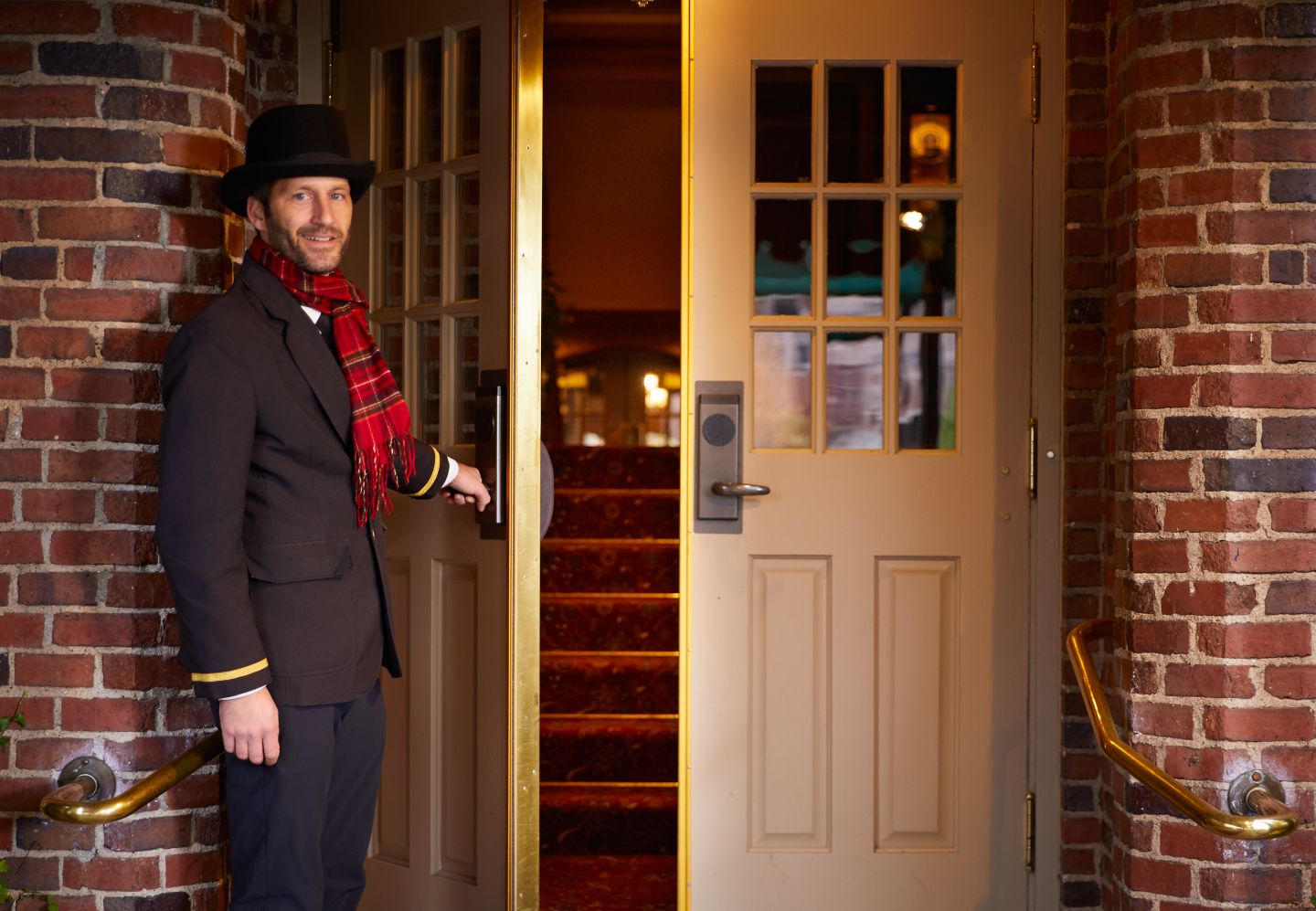 A bellman opens the entrance door of The American Club while smiling and wearing a red scarf