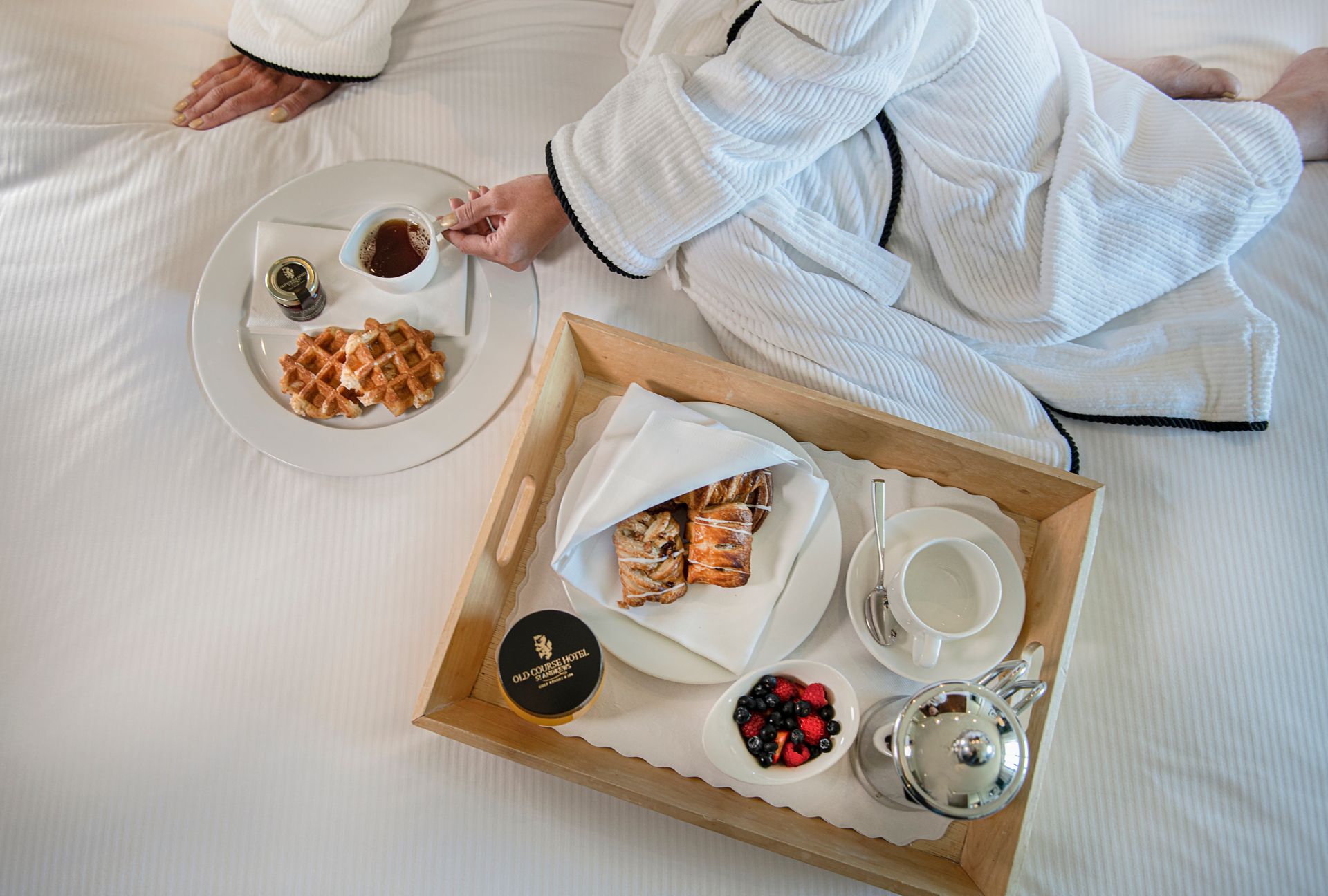 A guest sitting on the bed with a breakfast tray with syrup, waffles, pastry and fruit.