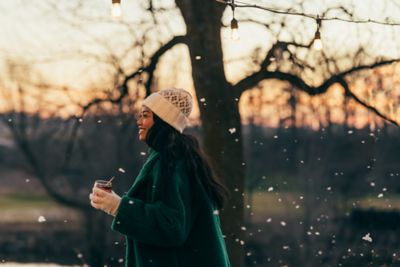 Smiling woman in a green coat and knit hat holding a drink outdoors at dusk, under string lights as snow falls.