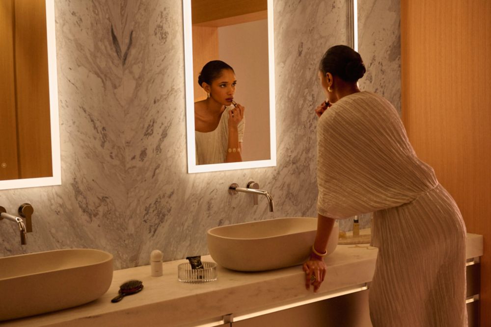 Woman in a white robe at a marble bathroom vanity with illuminated mirrors.