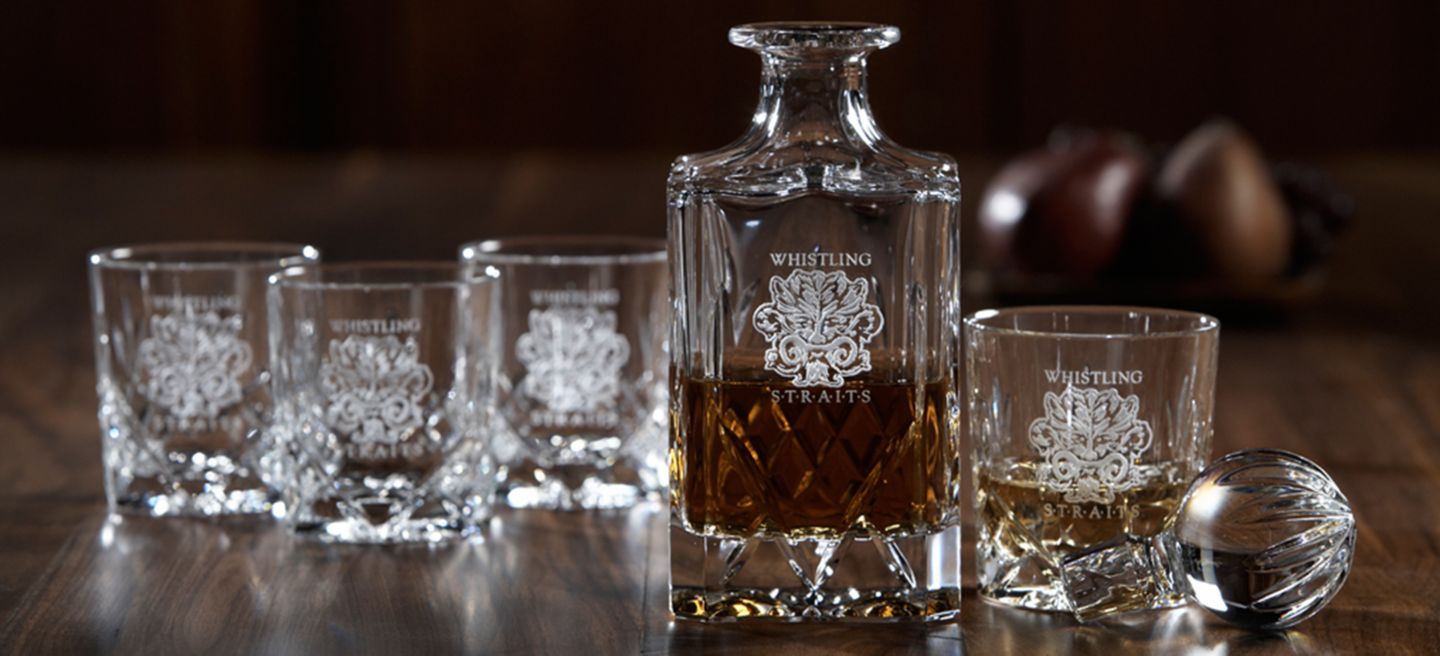A crystal decanter with brown alcohol featuring the Whistling Straits logos alongside four glasses with the Whistling Straits logo on them