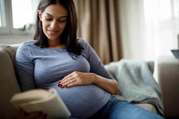 Pregnant woman sitting on a couch and she is reading a book