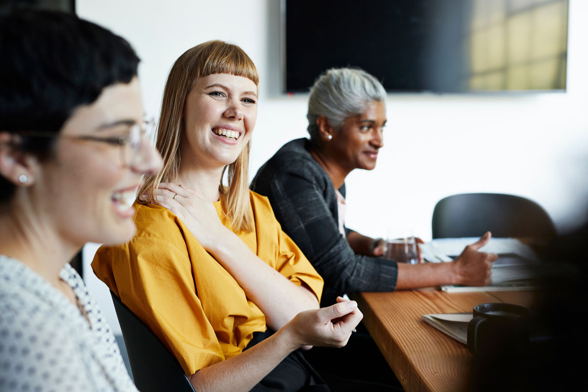 Three women sitting inside a meeting. The midde woman is the focus. She is smiling off into the distance with her hand on her shoulder