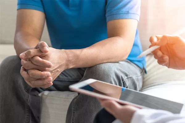 patient sitting down with his hands intertwined. There is a healthcare professional holding a tablet with a stylus