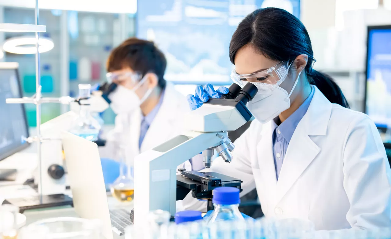 Two female scientists looking into microscopes insode of a lab. They are also wearing PPE