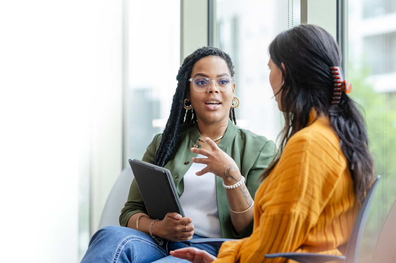 Two people having a sitting conversation. One is holding a tablet while taking with her hands while the other is sitting naturally