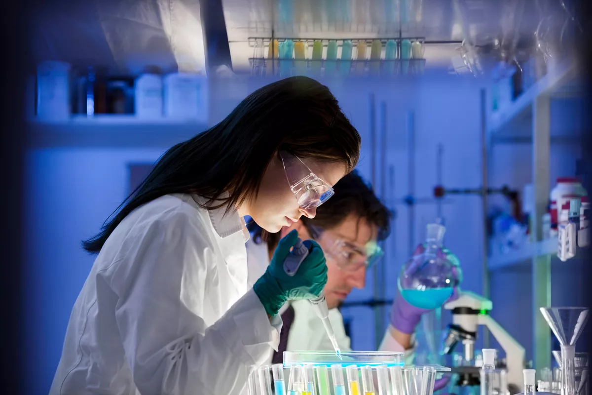 Two young scientists working hard inside of a lab. One is holding pipette, the other holding a beaker