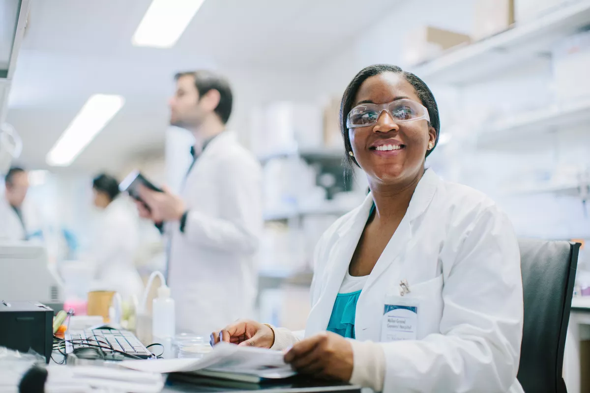 Scientist sitting at a desk in a lab. She is wearing goggles and has a big smile