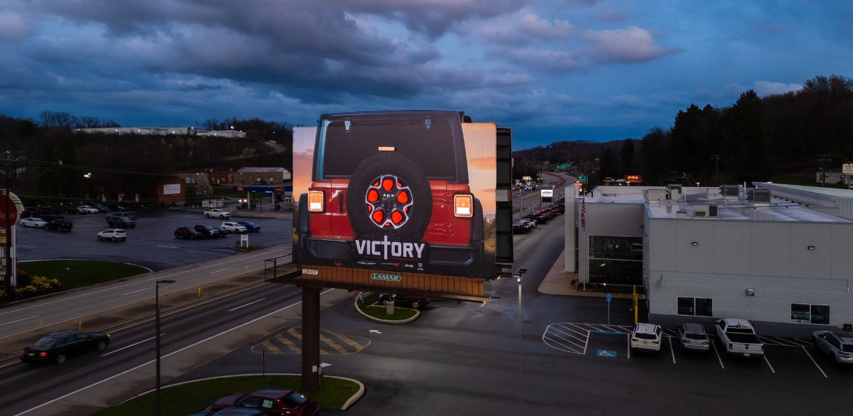 Lamar billboard featuring a Jeep Wrangler creative with an illuminated spare tire display beside a dealership at dusk