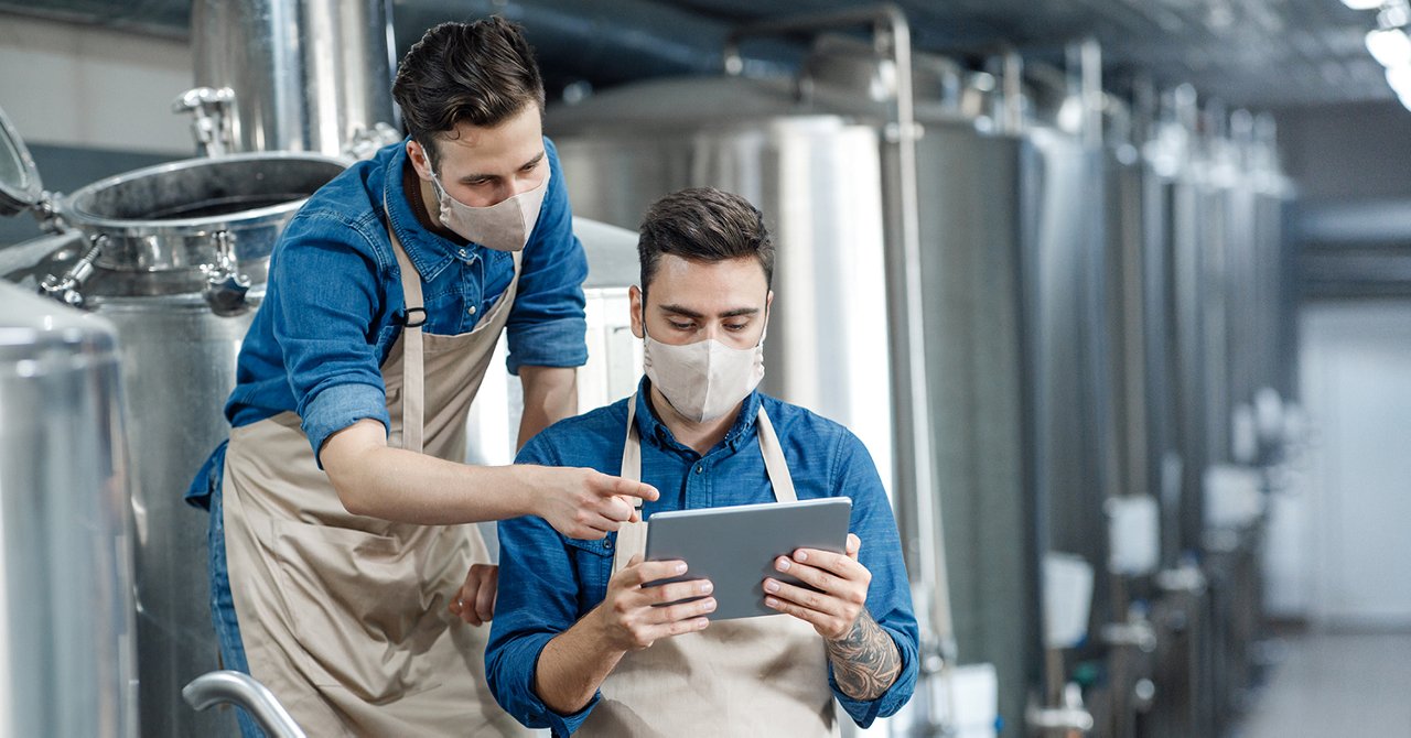Brewery workers enter data at tablet and operate equipment. Young guys in protective masks and aprons discuss and work with craft beer tanks in interior of factory during COVID-19 epidemic, free space; Shutterstock ID 1845133828; purchase_order: 3501043742; job: SPSD 2218; client: edostal@spotcreates.com; other: United States