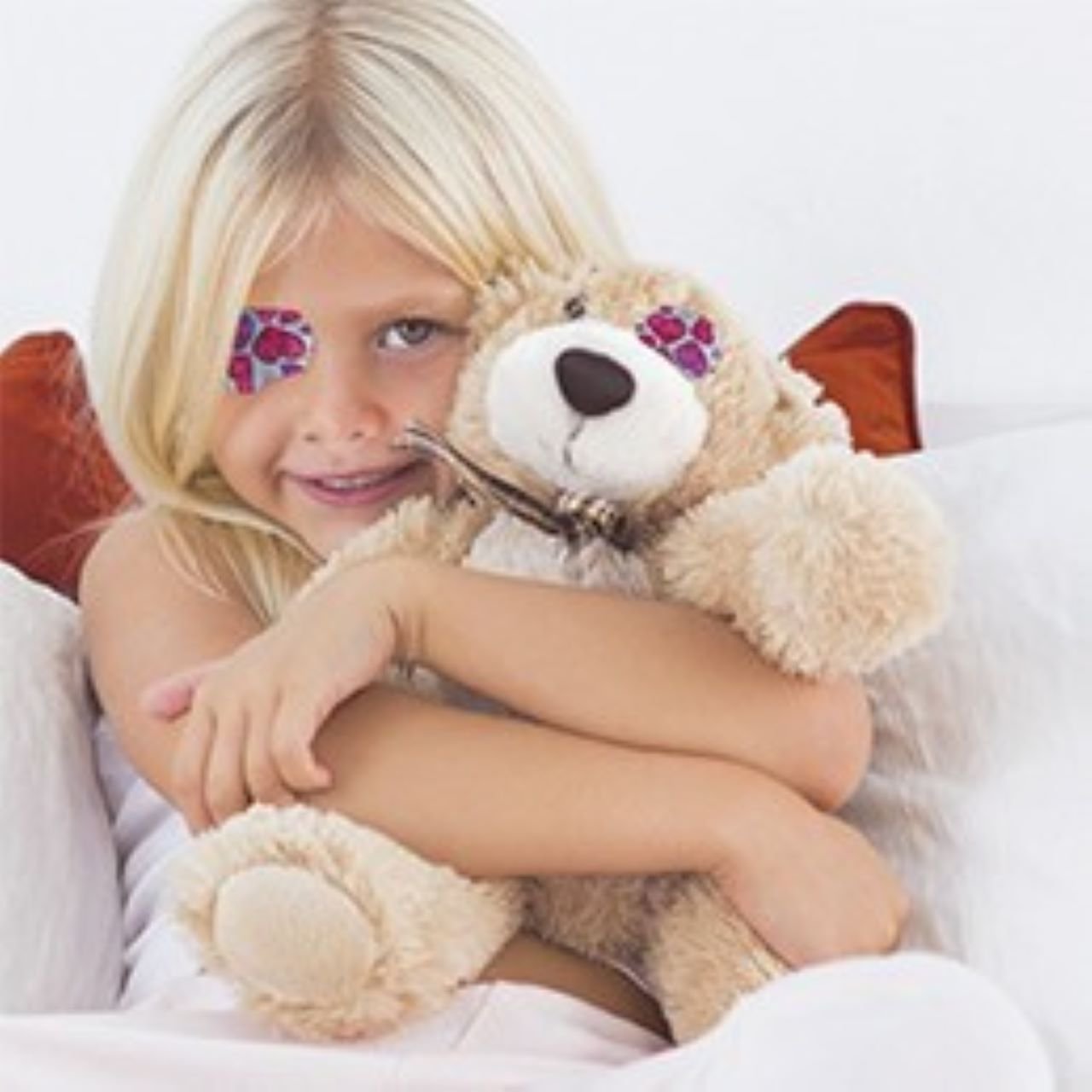 Blond meisje met Opticlude siliconen oogpleister met hartmotief, glimlachend en een beige-witte teddybeer omhelzend