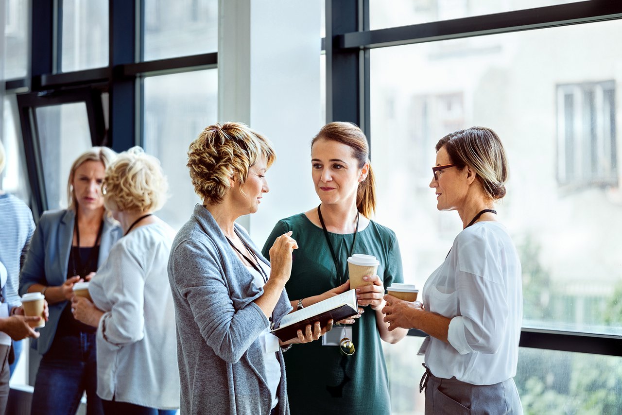 Three women talking to each other near a window with another small group of women talking in the background.