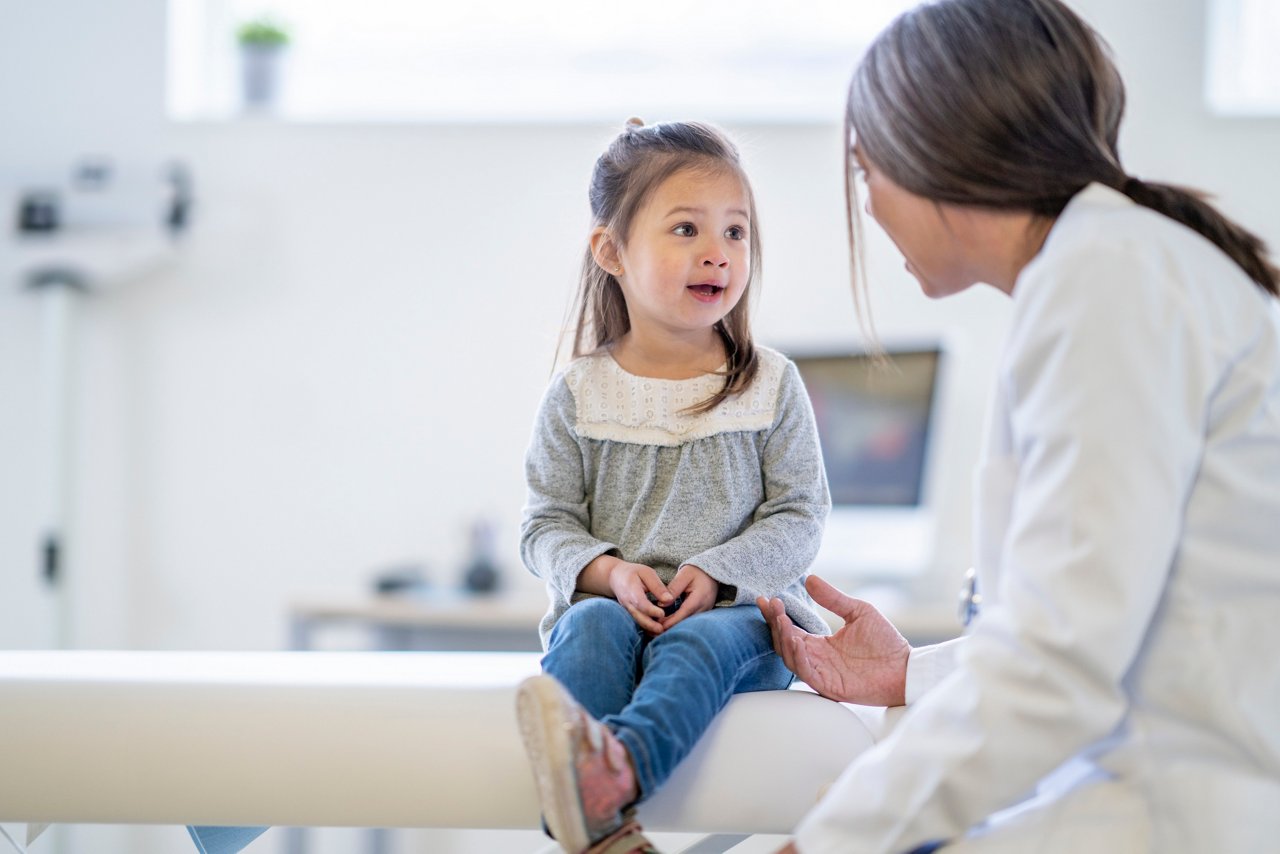 A young girl sits on the table in an examination room, excitedly talking to a doctor in a lab coat.