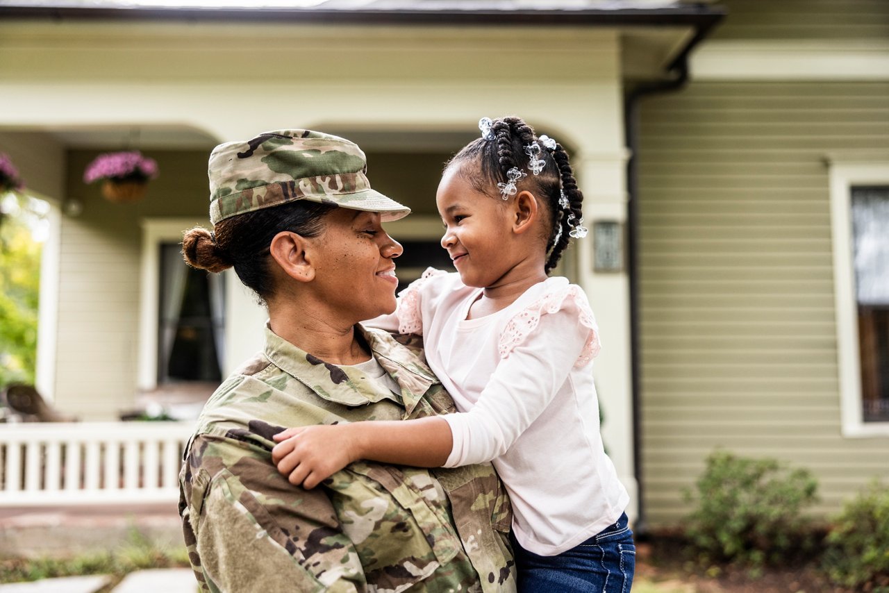 A woman in camouflage military fatigues holds her young daughter in her arms.