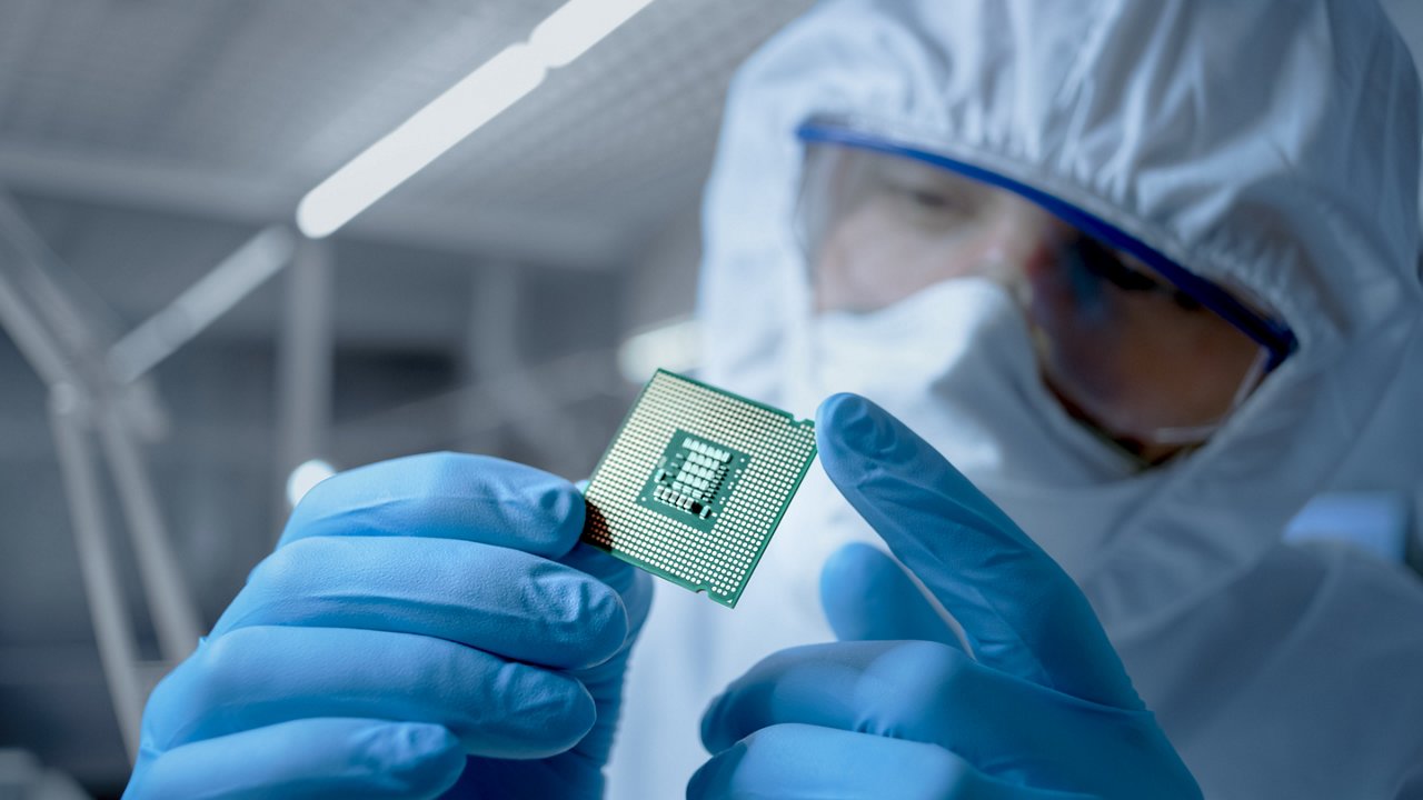 In Ultra Modern Electronic Manufacturing Factory Design Engineer in Sterile Coverall Holds Microchip with Gloves and Examines it.