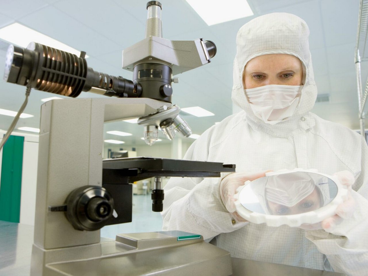 Woman in white clean room attire inspects a silicon wafer