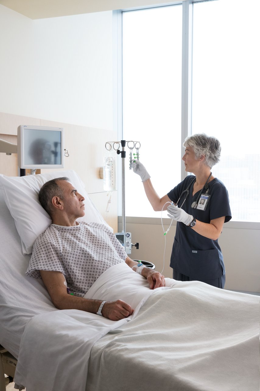 Nurse checking on male hospital patient who has PICC/CVC Securement Device + Tegaderm™ CHG Securement Dressing 1877-2100, Tegaderm™ CHG I.V. Securement Dressing 1660, and Curos™ disinfecting caps. Curos Tips™ Disinfecting Cap Strip for Male Luers - CM5-200 and Curos Jet™ Disinfecting Cap Strip for Needleless Connectors - CFJ5-250 hang on I.V. pole.