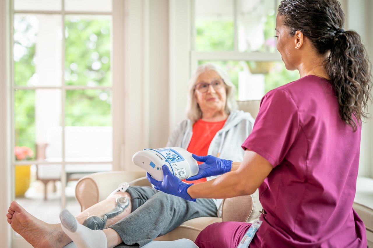 Patient in home with 3M™ Coban 2™ Two-Layer Compression System applied to wound. Nurse teaching patient how to use 3M™ ActiV.A.C.™ Therapy Unit.