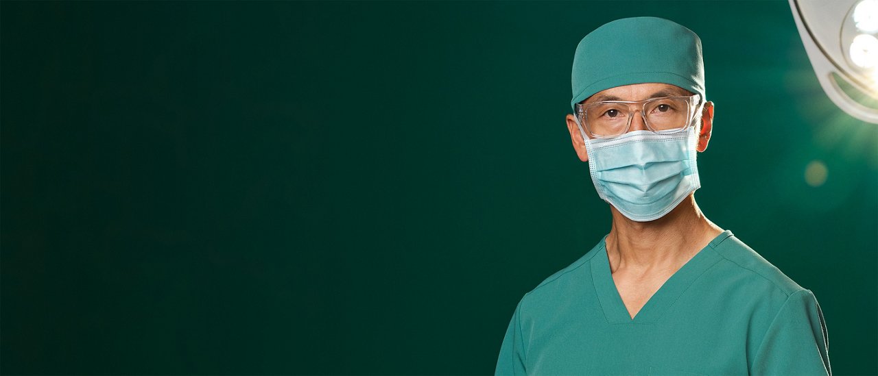 Standing OR male surgeon in teal scrubs and blue surgical mask with overhead OR light in background