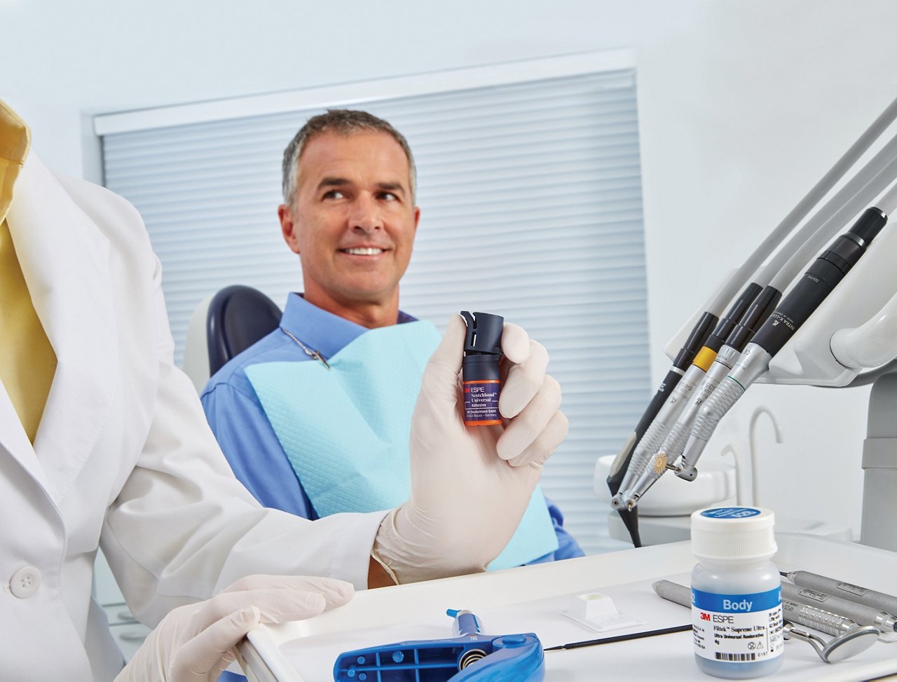 A patient seated in a dental chair is looking at the dental professional, who is holding a bottle of dental adhesive.  