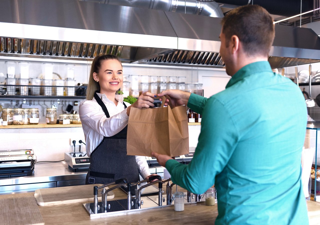 Cheerful waitress wearing apron serving customer at counter in restaurant - Small business and service concept with young business owner woman giving bag with takeaway food to client