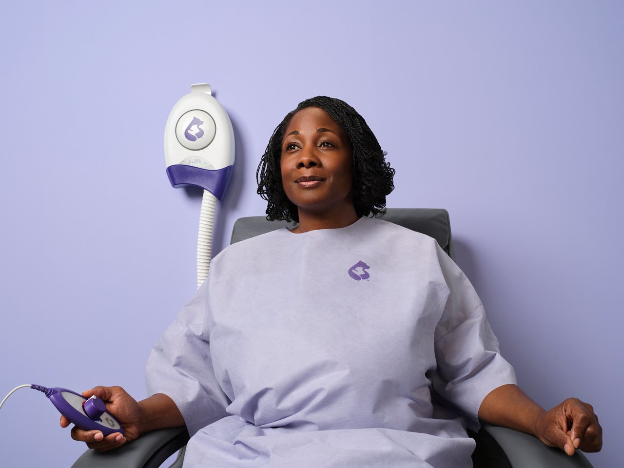 A female patient sits in a chair in a light purple room while wearing a 3M Bair Hugger Warming Gown System.