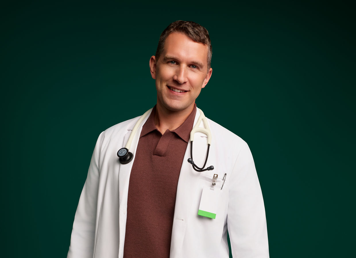 Porträt eines männlichen Arztes, stehend und lächelnd, mit Stethoskop um den Hals auf dunkelgrünem Hintergrund.