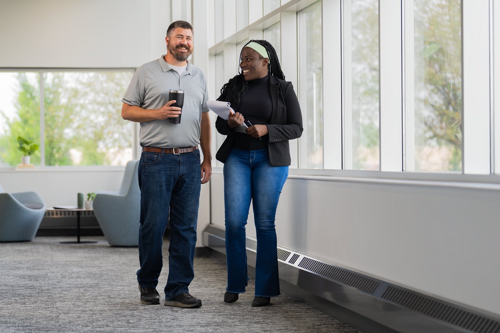 A man and woman employee talking in the hallway of an office building with large windows.