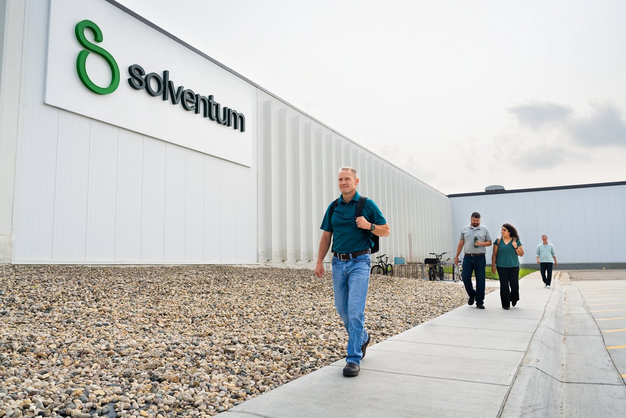 Four employees walk on a sidewalk outside in a staggered formation next to a white building with Solventum signage, full logo lockup..
