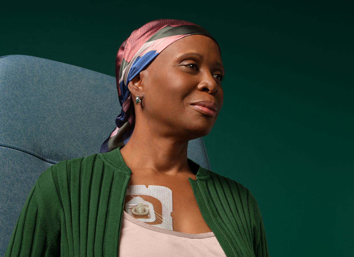 Adult female cancer patient sitting in hospital chair with IV port on chest covered by a Tegaderm dressing, on dark green background.