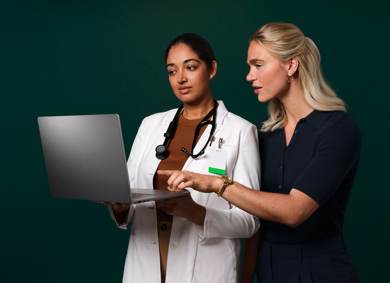 A physician wearing a white lab coat and stethoscope holds a laptop computer while another woman in business clothes points at the laptop and talks.
