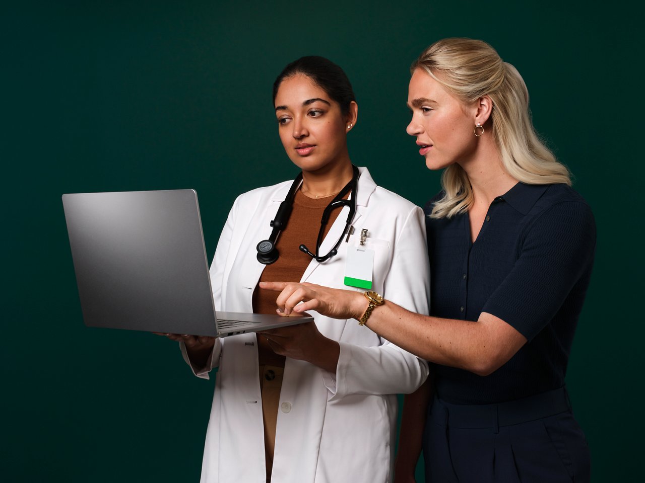 On a dark green background, a woman physician wearing a white lab coat and stethoscope holds a laptop computer while another woman in business clothes points at the laptop and talks.