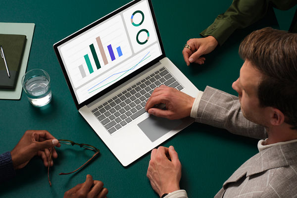 A laptop computer with colourful graphs and charts on screen sits on a dark green conference table where one man is controlling the laptop and two other people sit next to him.